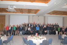 Geniş bir otel salonunda, ön tarafta yuvarlak masalar ve sandalyeler, arkada ise sahne önünde yan yana dizilmiş kalabalık bir grup görülüyor. Grup, resmi bir etkinlik kapsamında toplu fotoğraf için poz veriyor; salonun tavanı geometrik desenli ve aydınlatmalarla donatılmış.