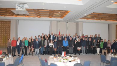 Geniş bir otel salonunda, ön tarafta yuvarlak masalar ve sandalyeler, arkada ise sahne önünde yan yana dizilmiş kalabalık bir grup görülüyor. Grup, resmi bir etkinlik kapsamında toplu fotoğraf için poz veriyor; salonun tavanı geometrik desenli ve aydınlatmalarla donatılmış.