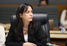 Bir toplantı salonunda oturan bir kadın, ciddi ve dikkatli bir ifadeyle konuşmaları dinlerken görülüyor.