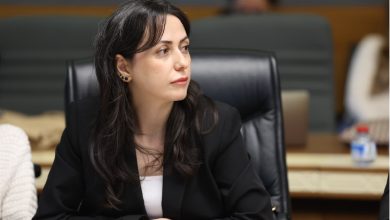 Bir toplantı salonunda oturan bir kadın, ciddi ve dikkatli bir ifadeyle konuşmaları dinlerken görülüyor.