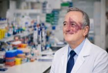 Laboratuvar ortamında, beyaz önlük ve kravatlı bir bilim insanı, arkasında deney malzemeleri bulunan tezgâh önünde otururken gülümseyerek kameranın dışına bakıyor. Yüzünün bir kısmında belirgin bir doğum lekesi görülüyor.