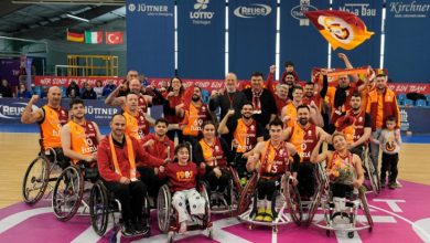 Spor salonunda, tekerlekli sandalyede oynayan basketbolcular ve destekçileri Galatasaray renkleri ve bayrağıyla birlikte sahada toplu halde zafer pozu vererek sevinçle kutlama yapıyor.