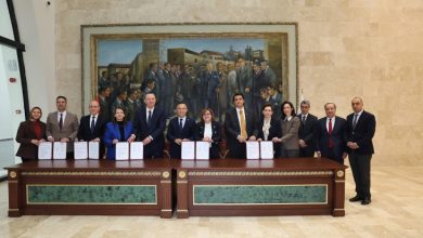 Protokol töreninde ayakta bulunan yetkililerin önünde uzun bir masa bulunmakta ve ellerinde tuttuklarını protokol dosyaları ile poz veriyorlar.