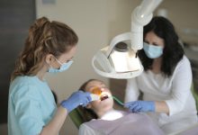 Bir diş kliniğinde hasta koltukta uzanmış şekilde tedavi görüyor. Maske ve eldiven takan iki sağlık çalışanı, güçlü bir muayene lambası altında hastanın ağzına müdahale ediyor. Ortam steril ve klinik bir atmosfer taşıyor.