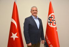 Görselde bir erkek resmi bir ortamda ayakta poz veriyor. Üzerinde lacivert bir blazer ceket ve açık renk gömlek var. Gülümseyerek kameraya bakıyor. Yanlarında iki bayrak bulunuyor: biri Türk bayrağı, diğeri ise kırmızı zemin üzerinde kurumsal bir logo ve yazı bulunan bir bayrak. Arka plan sade ve açık renkli bir duvardan oluşuyor.