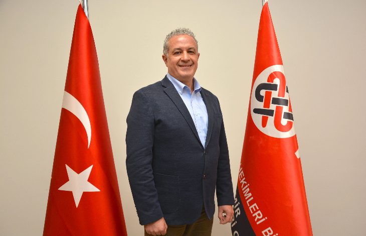 Görselde bir erkek resmi bir ortamda ayakta poz veriyor. Üzerinde lacivert bir blazer ceket ve açık renk gömlek var. Gülümseyerek kameraya bakıyor. Yanlarında iki bayrak bulunuyor: biri Türk bayrağı, diğeri ise kırmızı zemin üzerinde kurumsal bir logo ve yazı bulunan bir bayrak. Arka plan sade ve açık renkli bir duvardan oluşuyor.