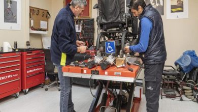 Bir atölyede iki görevli, tekerlekli sandalyeyi bakım ve onarım için çalışma tezgâhında tamir ediyor.