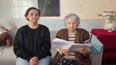 Fotoğrafta, ev ortamında yan yana oturan iki kadın görülüyor. Soldaki kadın kısa saçlı, siyah bir sweatshirt ve açık renk kot pantolon giymiş; ellerini dizlerinin üzerinde birleştirmiş şekilde kameraya doğru bakıyor. Sağdaki yaşlı kadın ise gri saçlı, desenli bir yelek ve çizgili bir kazak giymiş. Elinde birkaç sayfadan oluşan belgeler bulunuyor ve dikkatle bu kâğıtları okuyor.