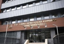 Çok katlı bir kamu binasının giriş cephesi görülüyor. Kapının üzerinde “T.C. Kamu Denetçiliği Kurumu (Ombudsmanlık)” yazısı yer alıyor. Binanın önünde demir parmaklıklar ve giriş merdivenleri bulunuyor. Resmi bir kurum binası izlenimi veren, dış mekânda çekilmiş bir fotoğraf.