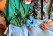 Bir ev ortamında sağlık çalışanı olduğu anlaşılan kişi, yeşil koruyucu önlük, maske ve eldivenle yaşlı bir kadının eline bakım uyguluyor. Yaşlı kadının üzeri battaniyeyle örtülü; işlem dikkatli ve özenli bir şekilde yapılıyor. Görsel, evde sağlık ve bakım hizmetini yansıtıyor.