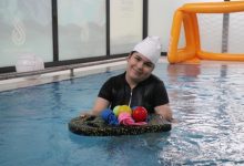 Havuzun içinde, bone takmış Zeynep Sema Taşdemir, gülümseyerek suyun üzerinde duran bir platformu tutuyor ve platformun üstünde renkli toplar yer alırken arka planda küçük bir su futbolu kalesi görünüyor.