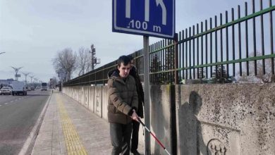 Kaldırımda beyaz bastonla ilerleyen görme engelli bir birey, yol kenarındaki engelleri fark etmeye çalışırken şehir trafiği arka planda görülüyor.
