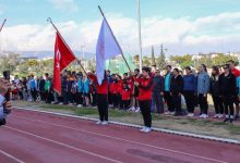 Bir atletizm pistinde tören düzenlenmiş. Önde iki sporcu Türk bayrağı ve organizasyon bayrağını tutuyor. Arkalarında sporcular ve katılımcılar sıra halinde dizilmiş, saygı duruşu ya da açılış anı gibi resmi bir atmosfer var.