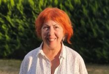 Açık havada, yeşil ağaçların önünde çekilmiş bir portre fotoğrafı. Kısa ve turuncu tonlarda saçlara sahip bir kadın, beyaz gömlekle kameraya doğru gülümseyerek poz veriyor. Gün ışığı yüzünü aydınlatıyor; arka plan doğal ve sakin bir ortam izlenimi veriyor.