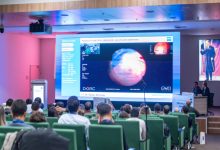 Görselde, bir konferans salonunda oturan izleyicilerin önünde büyük bir ekranda tıbbi bir sunum yapılırken, sahnede konuşmacıların retina görüntüleri içeren bir slaytı anlattığı görülüyor.