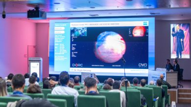 Görselde, bir konferans salonunda oturan izleyicilerin önünde büyük bir ekranda tıbbi bir sunum yapılırken, sahnede konuşmacıların retina görüntüleri içeren bir slaytı anlattığı görülüyor.