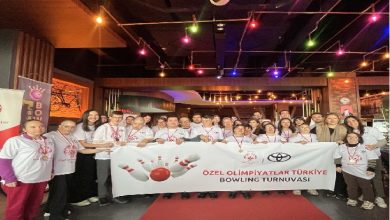 Kapalı bir mekânda kalabalık bir grup birlikte poz veriyor. Çoğu beyaz tişört giymiş ve boyunlarında madalyalar var. Önlerinde “Özel Olimpiyatlar Türkiye Bowling Turnuvası” yazılı bir pankart tutuluyor. Ortam renkli ışıklarla aydınlatılmış; eğlenceli ve kutlama havası taşıyan bir etkinlik görüntüsü veriyor.