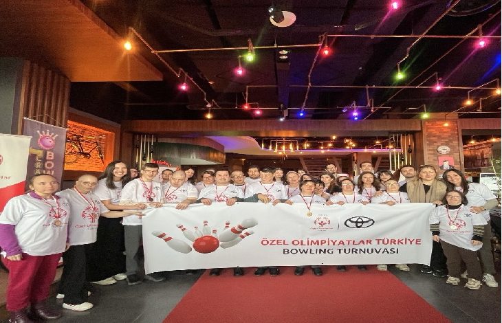 Kapalı bir mekânda kalabalık bir grup birlikte poz veriyor. Çoğu beyaz tişört giymiş ve boyunlarında madalyalar var. Önlerinde “Özel Olimpiyatlar Türkiye Bowling Turnuvası” yazılı bir pankart tutuluyor. Ortam renkli ışıklarla aydınlatılmış; eğlenceli ve kutlama havası taşıyan bir etkinlik görüntüsü veriyor.