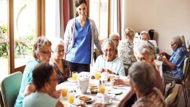 Aydınlık bir yemek salonunda, bir bakıcının eşliğinde masada birlikte kahvaltı yapan yaşlı bir grup insan görülüyor.