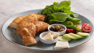 Kahvaltı tabağında sosyete simidi, karışık zeytin, domates, salatalık, üçgen krem peynir, beyaz peynir, roka yaprakları bulunuyor.