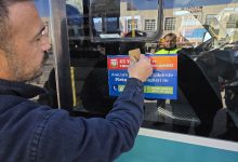 Bir kişi, şehir içi bir otobüsün camına bilgilendirici bir afiş yapıştırıyor. Camda yansıyan belediye görevlisi ve arka plandaki caddeyle birlikte sahne, toplu taşımada farkındalık ve denetim çalışmasını çağrıştırıyor.