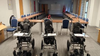 Bir toplantı salonunda U düzeninde yerleştirilmiş uzun masalar ve sandalyeler görülüyor. Salonun ön kısmında yan yana dizilmiş üç adet çocuklara yönelik özel tekerlekli sandalye/pozisyonlama cihazı bulunuyor. Arka tarafta projeksiyon perdesi ve Türk bayrakları yer alıyor. Ortam resmi bir tanıtım ya da teslim töreni havası veriyor.