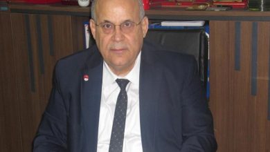 Bir ofiste, masa başında oturan takım elbiseli bir erkek görülüyor. Arkasında raflarda plaketler ve çeşitli ödüller yer alıyor; ortam resmi ve kurumsal bir çalışma alanını yansıtıyor.