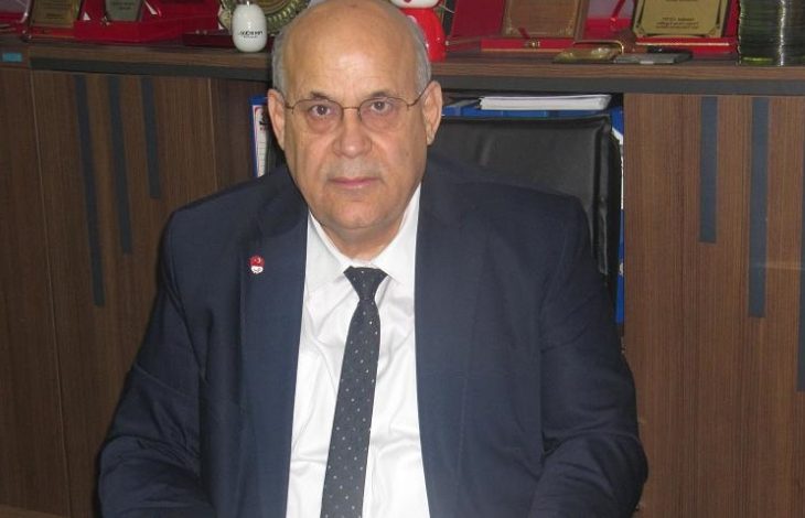 Bir ofiste, masa başında oturan takım elbiseli bir erkek görülüyor. Arkasında raflarda plaketler ve çeşitli ödüller yer alıyor; ortam resmi ve kurumsal bir çalışma alanını yansıtıyor.