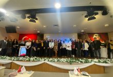 Bir konferans salonunda sahneye dizilmiş kalabalık bir grup toplu fotoğraf çektiriyor. Arkada Türk bayrağı ve Atatürk portresi yer alıyor. Ortada kürsü ve dijital ekran, sahne önünde beyaz çiçeklerle süsleme görülüyor. Resmî ve akademik bir etkinlik havası hakim.