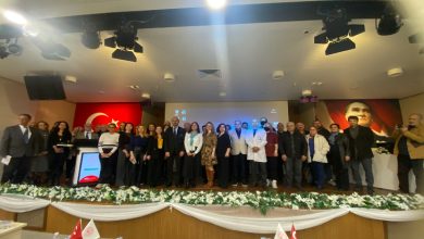 Bir konferans salonunda sahneye dizilmiş kalabalık bir grup toplu fotoğraf çektiriyor. Arkada Türk bayrağı ve Atatürk portresi yer alıyor. Ortada kürsü ve dijital ekran, sahne önünde beyaz çiçeklerle süsleme görülüyor. Resmî ve akademik bir etkinlik havası hakim.