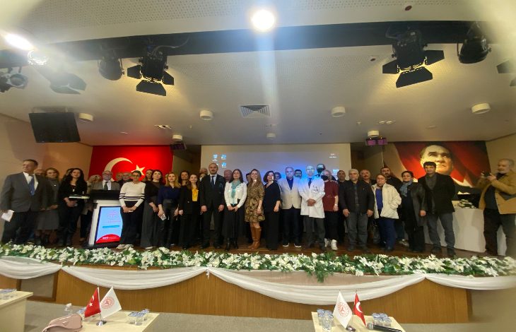 Bir konferans salonunda sahneye dizilmiş kalabalık bir grup toplu fotoğraf çektiriyor. Arkada Türk bayrağı ve Atatürk portresi yer alıyor. Ortada kürsü ve dijital ekran, sahne önünde beyaz çiçeklerle süsleme görülüyor. Resmî ve akademik bir etkinlik havası hakim.
