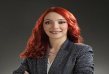 Görselde, profesyonel bir stüdyo ortamında çekilmiş bir portre fotoğrafı bulunuyor. Kırmızı saçlı, güler yüzlü bir kadın kamera karşısında poz veriyor. Üzerinde koyu renk bir blazer ceket ve açık renk bir bluz var. Kollarını hafifçe çaprazlayarak kendinden emin ve samimi bir duruş sergiliyor. Arka plan sade ve koyu tonlarda, bu da kişinin ön planda görünmesini sağlıyor.