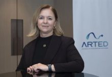 Görselde, kurumsal bir ortamda çekilmiş bir portre yer alıyor. Orta yaşlı bir kadın, siyah blazer ceket ve siyah üstle, bir masa ya da banko önünde poz vermiş. Arka planda sade bir ofis duvarı ve üzerinde “ARTED” yazılı bir logo bulunan bir pano görülüyor. Kadın kameraya doğrudan bakıyor ve hafif bir gülümsemeyle resmi bir duruş sergiliyor.