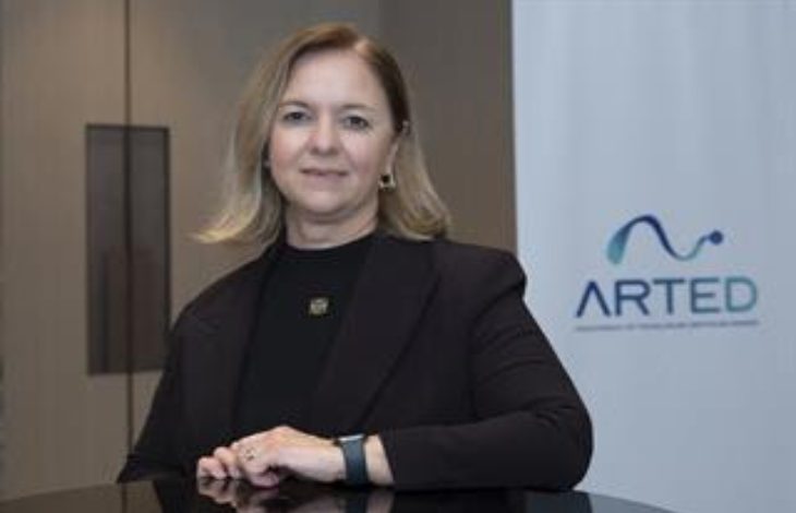 Görselde, kurumsal bir ortamda çekilmiş bir portre yer alıyor. Orta yaşlı bir kadın, siyah blazer ceket ve siyah üstle, bir masa ya da banko önünde poz vermiş. Arka planda sade bir ofis duvarı ve üzerinde “ARTED” yazılı bir logo bulunan bir pano görülüyor. Kadın kameraya doğrudan bakıyor ve hafif bir gülümsemeyle resmi bir duruş sergiliyor.