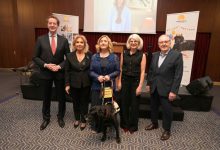 Bir konferans salonunda beş yetişkin yan yana poz veriyor. Ortadaki kadının yanında tasmalı siyah bir rehber köpek bulunuyor. Arkada sahne, projeksiyon ekranı ve etkinlik afişleri yer alıyor. Resmî ve anlamlı bir tören havası dikkat çekiyor.