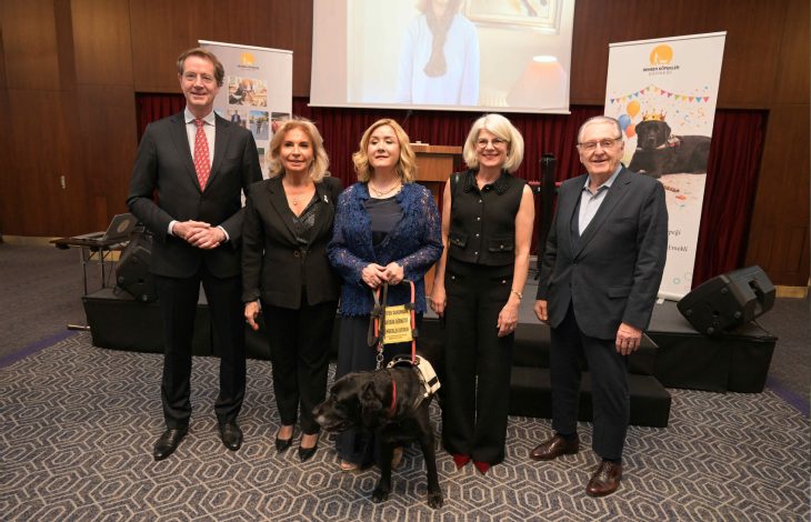 Bir konferans salonunda beş yetişkin yan yana poz veriyor. Ortadaki kadının yanında tasmalı siyah bir rehber köpek bulunuyor. Arkada sahne, projeksiyon ekranı ve etkinlik afişleri yer alıyor. Resmî ve anlamlı bir tören havası dikkat çekiyor.