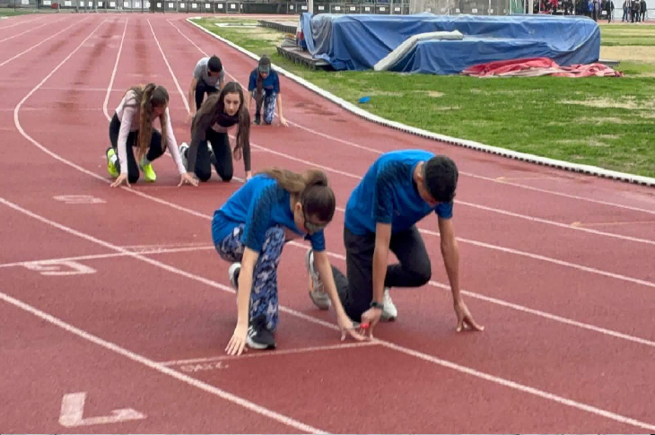 Atletizm pistinde genç sporcular старт pozisyonunda diz çökmüş halde bekliyor. Eller çizgide, gözler önde; start anına kilitlenmişler. Arka planda diğer sporcular ve saha ekipmanları görünüyor; yarışın heyecanı saniyelerle ölçülüyor.