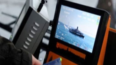 Bir toplu taşıma aracında yer alan elektronik kart okuma cihazı görülüyor. Ekranda deniz üzerinde ilerleyen bir vapur görüntüsü yer alıyor. Ön planda bir kişi ulaşım kartını cihaza tutuyor. Görsel, toplu taşımada kartlı geçiş sistemini ve elektronik bilet uygulamasını yansıtıyor.