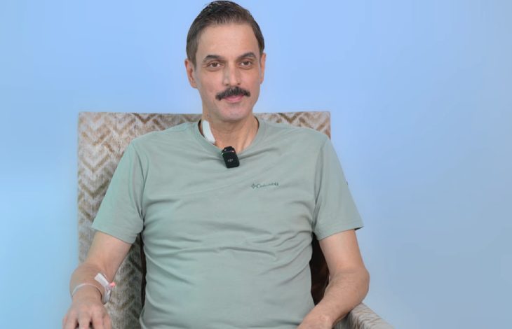 Açık mavi arka plan önünde bir koltukta oturan, oyuncu Ufuk Özkan açık yeşil tişört giymiş ve yakasında küçük bir mikrofon takılı şekilde, kolunda ve boynunda damar yolu pansumanları bulunarak kameraya doğru bakıyor.