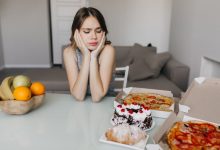 Masada oturan genç bir kadın, elleriyle yüzünü tutmuş halde kararsız ve stresli görünüyor. Önünde pizza ve tatlılar, yanında ise meyveler bulunuyor. Görsel, sağlıksız ve sağlıklı beslenme arasındaki ikilemi yansıtıyor.