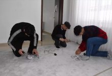 Bir odada üç genç yerde diz çökmüş şekilde elektrik kabloları ve küçük cihazlarla uğraşıyor. Önlerinde uzatma kablosu ve bağlantı aparatları bulunuyor. Oda sade; beyaz duvarlar ve tül perde arka planda görülüyor. Gençler bir cihazın kurulumunu ya da bağlantısını yapıyor gibi görünüyor.