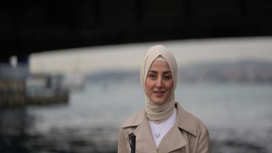 Görme engelli sosyolog ve kamu çalışanı Esra Durmuş, arka planda deniz ve iskele silüeti bulanık şekilde görünürken kameraya doğru hafif bir gülümsemeyle bakıyor.