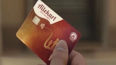 Fotoğrafta bir kişinin elinde kırmızı tonlarında tasarlanmış temassız özellikli bir AileKart tutuluyor. Kartın üzerinde aile figürü simgesi ve çip bulunurken arka plan bulanık şekilde bir bina girişini andırıyor.