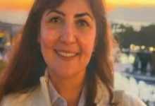 Gün batımı ışığında çekilmiş bir fotoğrafta, uzun koyu renk saçlı bir kadın kameraya gülümseyerek bakıyor. Açık renk bir ceket ve gömlek giymiş. Arka planda su kenarı, ağaçlar ve loş şehir ışıkları görülüyor; sıcak tonlu, sakin bir atmosfer hâkim.