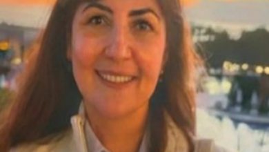 Gün batımı ışığında çekilmiş bir fotoğrafta, uzun koyu renk saçlı bir kadın kameraya gülümseyerek bakıyor. Açık renk bir ceket ve gömlek giymiş. Arka planda su kenarı, ağaçlar ve loş şehir ışıkları görülüyor; sıcak tonlu, sakin bir atmosfer hâkim.