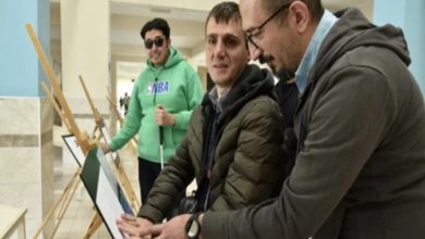 Bir üniversite koridorunda üç kişi kabartma bir tabloyu inceliyor. Ortadaki görme engelli birey, eserin yüzeyine dokunarak kabartma detayları hissederken yanında bulunan kişi ona rehberlik ediyor. Arka planda ise elinde beyaz baston bulunan başka bir görme engelli birey ve sergileme panoları görülüyor.