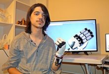 Bir odada, omuz hizasında uzun saçlı genç bir kişi kameraya bakıyor. Kolunda kabloları görünen mekanik bir protez el takılı. Arka planda bir masanın üzerinde bilgisayar ekranı var; ekranda robotik bir elin teknik çizimi ya da 3D modeli görünüyor. Ortam, teknoloji ve mühendislik çalışması yapılan bir atölye havası veriyor.