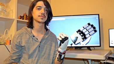 Bir odada, omuz hizasında uzun saçlı genç bir kişi kameraya bakıyor. Kolunda kabloları görünen mekanik bir protez el takılı. Arka planda bir masanın üzerinde bilgisayar ekranı var; ekranda robotik bir elin teknik çizimi ya da 3D modeli görünüyor. Ortam, teknoloji ve mühendislik çalışması yapılan bir atölye havası veriyor.