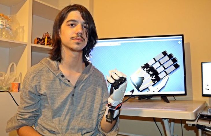 Bir odada, omuz hizasında uzun saçlı genç bir kişi kameraya bakıyor. Kolunda kabloları görünen mekanik bir protez el takılı. Arka planda bir masanın üzerinde bilgisayar ekranı var; ekranda robotik bir elin teknik çizimi ya da 3D modeli görünüyor. Ortam, teknoloji ve mühendislik çalışması yapılan bir atölye havası veriyor.