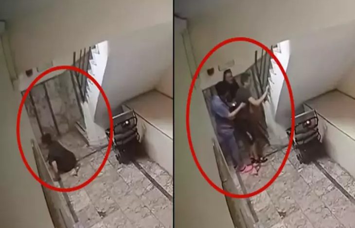 Görsel, iki ayrı güvenlik kamerası karesinden oluşuyor. Her iki karede de kırmızı bir daire içinde merdiven boşluğu görülüyor. İlk karede bir kişinin merdivenlerde oturur ya da çömelir halde olduğu dikkati çekiyor. İkinci karede ise aynı noktada üç kişinin bir arada durduğu, ortadaki kişiye destek verildiği anlaşılıyor. Merdiven kenarında bir engelli asansörü veya taşıma cihazı bulunduğu görülüyor. Kareler, kapalı bir apartman içi koridorunda kaydedilmiş izlenimi veriyor.